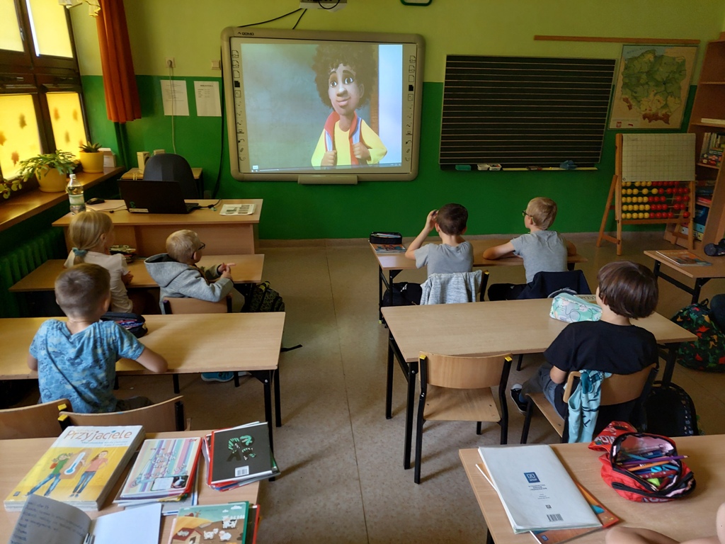 Klasa szkolna. Uczniowie siedzą w ławkach i oglądają bajkę pod tytułem "Jak zostałem bohaterem".