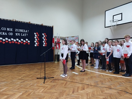 Udekorowana sala gimnastyczna. Na środku stoją uczniowie ubrani w strój galowy. Recytują wiersze i śpiewają pieśni.