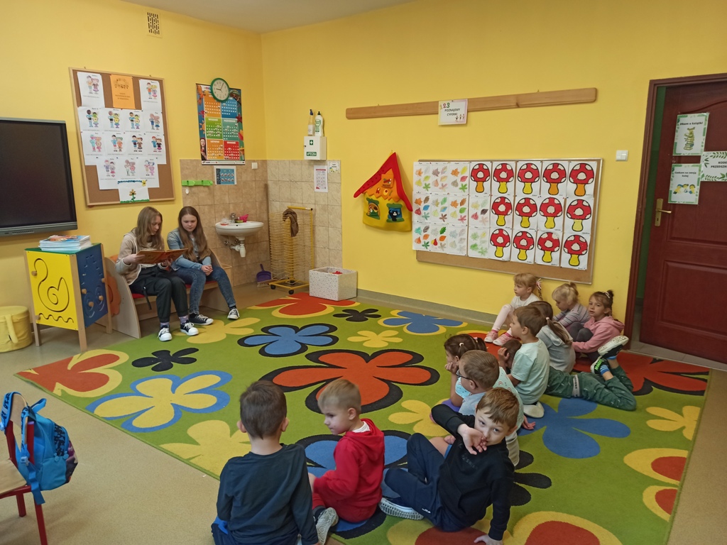 Sala przedszkolna. Na dywanie siedzą przedszkolaki. Przed nimi na krzesłach siedzą dwie uczennice. Czytają głośno przedszkolakom książkę.