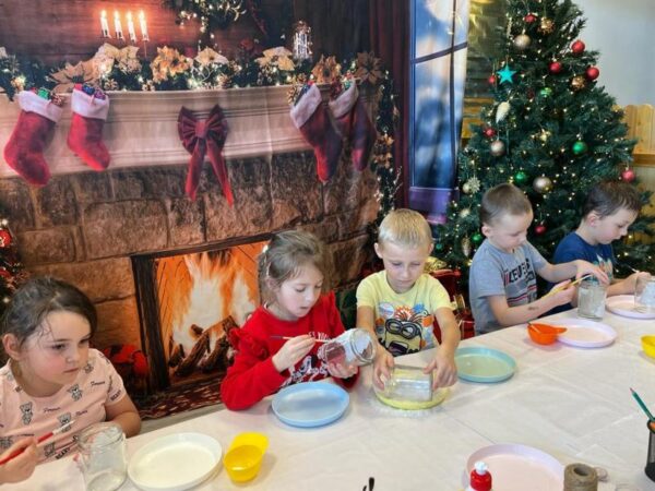 Dzieci siedzą dookoła stołu. Wykonują świąteczne stroiki ze słoików, papieru i innych materiałów.