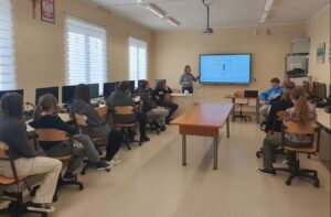 Dziesięciu uczniów klasy siódmej podczas lekcji w sali konputerowej obserwuje prezentowane materiały na tablicy interaktywnej przez wychowawczynię