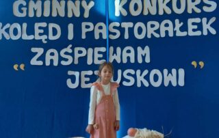 Na środku sali stoi dziewczynka i śpiewa kolędę. Za nią znajduje się niebieskie tło z napisem Gminny Konkurs Kolęd i Pastorałek "Zaśpiewam Jezuskowi".