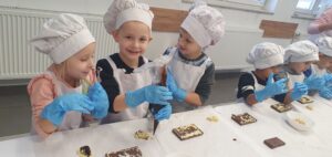 grupa dzieci w czapkach kucharskich, fartuchach i rękawicach wykonujące słodycze