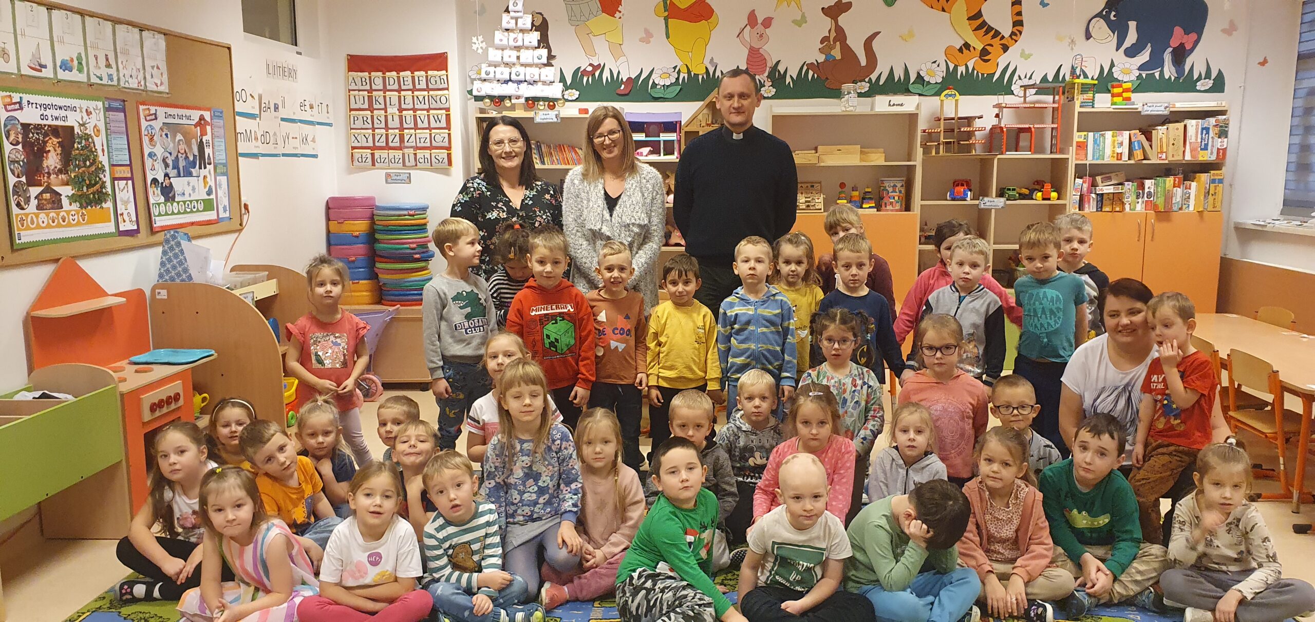 grupa dzieci w sali przedszkolnej wraz z opiekunami i księdzem