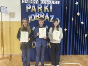 Trzech uczniów stoi na sali gimnastycznej i prezentuje dyplomy oraz nagrody otrzymane w konkursie