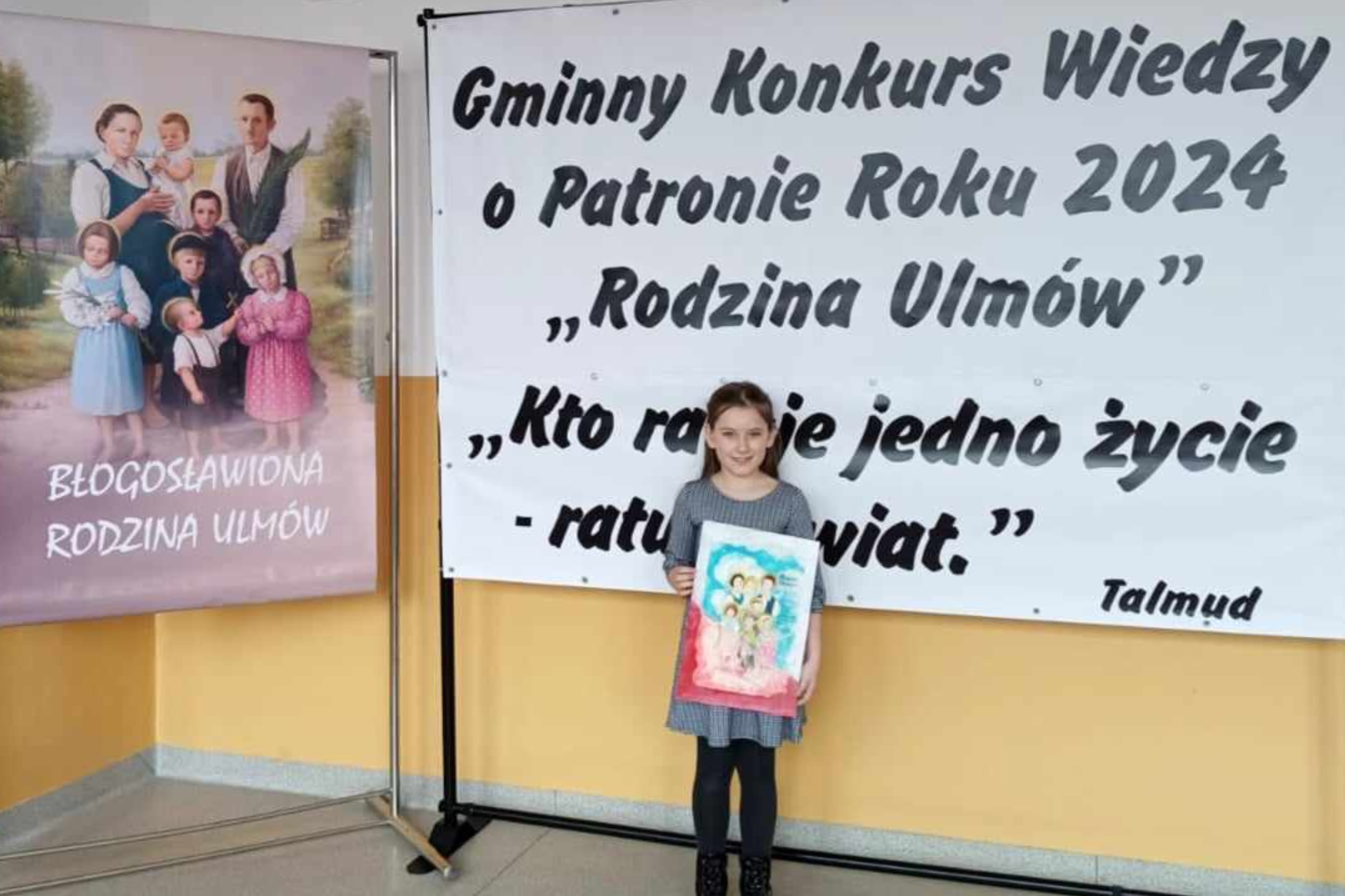 Uczennica klasy I stoi przez plakatem konkursu Patron Roku 2024 i prezentuje otrzymaną nagrodę.