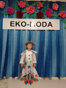 Dziewczynka prezentuje strój przygotowany na konkurs. W tle napis Eko-Moda oraz czerwone kwiaty na niebieskim płótnie.