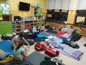 Uczniowie klasy czwartej siedzą w swojej klasie. Kilkoro gra w bierki, inni rozmawiają i przygotowują się do snu.