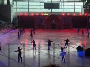 Siedemnastu łyżwiarzy tańczy na lodzie. W tle scenografia do rewii.