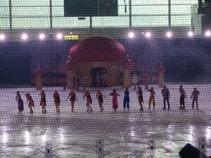 Piętnastu łyżwiarzy tańczy na lodzie. W tle scenografia do rewii.