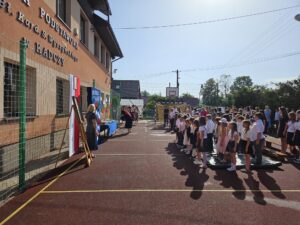 Dyrektor szkoły i grupa młodzieży na boisku szkolnym przed szkołą podczas zakończenia roku szkolnego