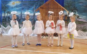 na obrazku widzimy grupę dzieci w białych sukienkach. W tle widzimy stajenkę i zimową scenerię