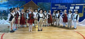 na obrazku widzimy grupę dzieci w strojach kolędniczych siedzących . W tle widzimy stajenkę i zimową scenerię