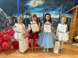 obrazek przedstawia cztery dziewczynki w strojach kolędniczych trzymające z dyplomami ni prezentami. W tle mamy bożonarodzeniową scenerię