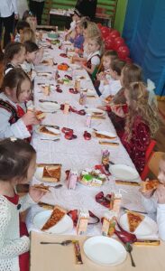 obrazek przedstawia grupę dzieci siedzących przy stole. Na stole są talerzyki z pizzą, soczki i słodkie przekąski