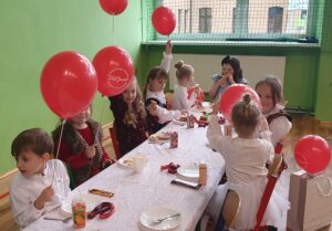 obrazek przedstawia grupę dzieci siedzących przy stole. Na stole są talerzyki z pizzą, soczki i słodkie przekąski. Dzieci trzymają w ręce czerwone balony