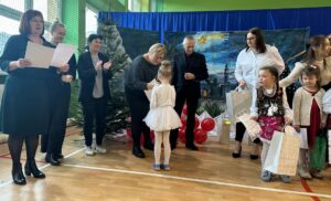 obrazek przedstawia grupę ludzi dorosłych i dzieci na tle bożonarodzeniowej dekoracji.  Dorośli trzymają w ręce kartki, a dzieci prezenty
