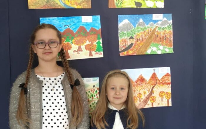 Zdjęcie przedstawia dwie dziewczynki stojące na tle wystawy prac plastycznych o tematyce górskiej. Nad rysunkami widnieje cytat zachęcający do poznawania własnych gór. Obok znajdują się papierowe torby, prawdopodobnie z nagrodami. Dziewczynki są elegancko ubrane, co sugeruje udział w konkursie lub uroczystości związanej z prezentacją prac.