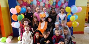 Zdjęcie przedstawia grupę dzieci w kolorowych strojach karnawałowych, pozujących na tle dekoracji z balonami. Dzieci przebrane są za różne postacie, takie jak Darth Vader, szturmowiec, czarodziej, superbohater, papuga, kotek czy piłkarz. Stroje są pełne kreatywności, a atmosfera jest radosna i pełna zabawy.