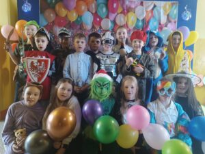 Zdjęcie przedstawia grupę dzieci w kolorowych strojach karnawałowych, pozujących na tle dekoracji z balonami. Dzieci przebrane są za różne postacie, takie jak Darth Vader, szturmowiec, czarodziej, superbohater, papuga, kotek czy piłkarz. Stroje są pełne kreatywności, a atmosfera jest radosna i pełna zabawy.