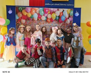 Zdjęcie przedstawia grupę dzieci w kolorowych strojach karnawałowych, pozujących na tle dekoracji z balonami. Dzieci przebrane są za różne postacie, takie jak Darth Vader, szturmowiec, czarodziej, superbohater, papuga, kotek czy piłkarz. Stroje są pełne kreatywności, a atmosfera jest radosna i pełna zabawy.