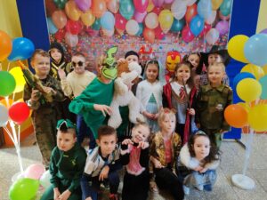 Zdjęcie przedstawia grupę dzieci w kolorowych strojach karnawałowych, pozujących na tle dekoracji z balonami. Dzieci przebrane są za różne postacie, takie jak Darth Vader, szturmowiec, czarodziej, superbohater, papuga, kotek czy piłkarz. Stroje są pełne kreatywności, a atmosfera jest radosna i pełna zabawy.