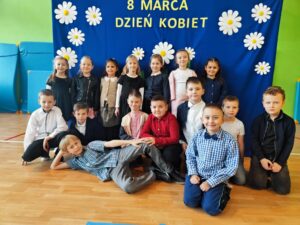 Zdjęcie przedstawia grupę dzieci pozujących na tle niebieskiego banera ozdobionego białymi kwiatami i napisem „8 MARCA DZIEŃ KOBIET”. Dzieci są ubrane elegancko – dziewczynki w sukienkach lub spódnicach, a chłopcy w koszulach, niektórzy z muszkami. Są uśmiechnięte i ustawione w kilku rzędach – część stoi, część klęczy, a jedno z dzieci leży na podłodze w swobodnej pozie. Zdjęcie zostało wykonane w sali gimnastycznej lub szkolnej świetlicy, co widać po materacach na ścianach