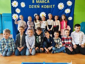 Zdjęcie przedstawia grupę dzieci pozujących na tle niebieskiego banera ozdobionego białymi kwiatami i napisem „8 MARCA DZIEŃ KOBIET”. Dzieci są ubrane elegancko – dziewczynki w sukienkach lub spódnicach, a chłopcy w koszulach, niektórzy z muszkami. Są uśmiechnięte i ustawione w kilku rzędach – część stoi, część klęczy, a jedno z dzieci leży na podłodze w swobodnej pozie. Zdjęcie zostało wykonane w sali gimnastycznej lub szkolnej świetlicy, co widać po materacach na ścianach