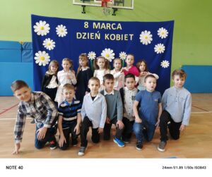 Zdjęcie przedstawia grupę dzieci pozujących na tle niebieskiego banera ozdobionego białymi kwiatami i napisem „8 MARCA DZIEŃ KOBIET”. Dzieci są ubrane elegancko – dziewczynki w sukienkach lub spódnicach, a chłopcy w koszulach, niektórzy z muszkami. Są uśmiechnięte i ustawione w kilku rzędach – część stoi, część klęczy, a jedno z dzieci leży na podłodze w swobodnej pozie. Zdjęcie zostało wykonane w sali gimnastycznej lub szkolnej świetlicy, co widać po materacach na ścianach