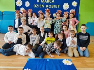 Zdjęcie przedstawia grupę dzieci pozujących na tle niebieskiego banera ozdobionego białymi kwiatami i napisem „8 MARCA DZIEŃ KOBIET”. Dzieci są ubrane elegancko – dziewczynki w sukienkach lub spódnicach, a chłopcy w koszulach, niektórzy z muszkami. Są uśmiechnięte i ustawione w kilku rzędach – część stoi, część klęczy, a jedno z dzieci leży na podłodze w swobodnej pozie. Zdjęcie zostało wykonane w sali gimnastycznej lub szkolnej świetlicy, co widać po materacach na ścianach