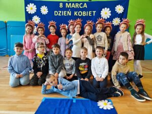 Zdjęcie przedstawia grupę dzieci pozujących na tle niebieskiego banera ozdobionego białymi kwiatami i napisem „8 MARCA DZIEŃ KOBIET”. Dzieci są ubrane elegancko – dziewczynki w sukienkach lub spódnicach, a chłopcy w koszulach, niektórzy z muszkami. Są uśmiechnięte i ustawione w kilku rzędach – część stoi, część klęczy, a jedno z dzieci leży na podłodze w swobodnej pozie. Zdjęcie zostało wykonane w sali gimnastycznej lub szkolnej świetlicy, co widać po materacach na ścianach