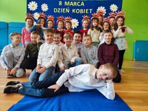Zdjęcie przedstawia grupę dzieci pozujących na tle niebieskiego banera ozdobionego białymi kwiatami i napisem „8 MARCA DZIEŃ KOBIET”. Dzieci są ubrane elegancko – dziewczynki w sukienkach lub spódnicach, a chłopcy w koszulach, niektórzy z muszkami. Są uśmiechnięte i ustawione w kilku rzędach – część stoi, część klęczy, a jedno z dzieci leży na podłodze w swobodnej pozie. Zdjęcie zostało wykonane w sali gimnastycznej lub szkolnej świetlicy, co widać po materacach na ścianach