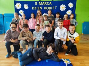 Zdjęcie przedstawia grupę dzieci pozujących na tle niebieskiego banera ozdobionego białymi kwiatami i napisem „8 MARCA DZIEŃ KOBIET”. Dzieci są ubrane elegancko – dziewczynki w sukienkach lub spódnicach, a chłopcy w koszulach, niektórzy z muszkami. Są uśmiechnięte i ustawione w kilku rzędach – część stoi, część klęczy, a jedno z dzieci leży na podłodze w swobodnej pozie. Zdjęcie zostało wykonane w sali gimnastycznej lub szkolnej świetlicy, co widać po materacach na ścianach