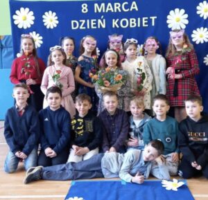 Zdjęcie przedstawia grupę dzieci pozujących na tle niebieskiego banera ozdobionego białymi kwiatami i napisem „8 MARCA DZIEŃ KOBIET”. Dzieci są ubrane elegancko – dziewczynki w sukienkach lub spódnicach, a chłopcy w koszulach, niektórzy z muszkami. Są uśmiechnięte i ustawione w kilku rzędach – część stoi, część klęczy, a jedno z dzieci leży na podłodze w swobodnej pozie. Zdjęcie zostało wykonane w sali gimnastycznej lub szkolnej świetlicy, co widać po materacach na ścianach
