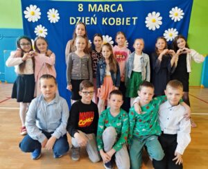 Zdjęcie przedstawia grupę dzieci pozujących na tle niebieskiego banera ozdobionego białymi kwiatami i napisem „8 MARCA DZIEŃ KOBIET”. Dzieci są ubrane elegancko – dziewczynki w sukienkach lub spódnicach, a chłopcy w koszulach, niektórzy z muszkami. Są uśmiechnięte i ustawione w kilku rzędach – część stoi, część klęczy, a jedno z dzieci leży na podłodze w swobodnej pozie. Zdjęcie zostało wykonane w sali gimnastycznej lub szkolnej świetlicy, co widać po materacach na ścianach