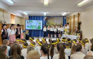 Rozpoczęcie akademii. Po lewej stronie 6 uczniów klasy 4 w strojach regionalnych. Na froncie monitor, na którym wyświetlana była prezentacja oraz uczniowie klasy 5 na tle dekoracyjnego płótna (kwitnące drzewo, pisanki). Po prawej wielkanocny stół.