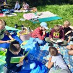 Uczniowie siedzą na kocach na zielonym placu za szkołą i czytają książki