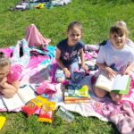Trzy uczennice siedzą na kocach na zielonym placu za szkołą i czytają książki