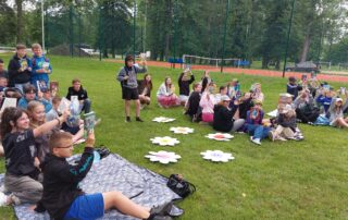 Uczniowie na zielonym placu, siedzą na kocach lub karimatach i pokazują ulubione książki