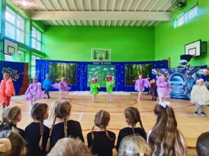 Grupa dzieci w kolorowych kostiumach tańczy na sali gimnastycznej podczas przedstawienia szkolnego zatytułowanego 'Moja Planeta'. Scenografia w tle przedstawia las, Ziemię oraz futurystyczną stację kosmiczną. Widzowie – inne dzieci – siedzą z przodu, obserwując występ