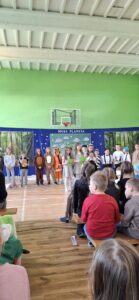 Grupa dzieci z klasy 2 stoi na scenie w sali gimnastycznej, biorąc udział w przedstawieniu pt. 'Moja Planeta'. Część dzieci przebrana jest za zwierzęta leśne, inne mają odświętne stroje lub tematyczne koszulki. Dwoje dzieci trzyma zielone kartki i mówi do mikrofonu. Przed sceną siedzi publiczność złożona z uczniów.