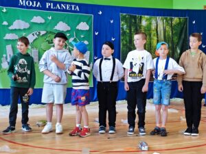 Grupa siedmiu chłopców z klasy 2 stoi na sali gimnastycznej podczas szkolnego występu. W tle znajduje się dekoracja z napisem 'MOJA PLANETA', przedstawiająca zieloną Ziemię i las. Dzieci są kolorowo ubrane, niektórzy mają czapki z daszkiem, a dwóch chłopców nosi koszulki z Pikachu. Na podłodze widoczne są rekwizyty z występu