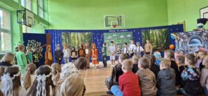 „Grupa dzieci w kolorowych kostiumach tańczy na sali gimnastycznej podczas przedstawienia szkolnego zatytułowanego 'Moja Planeta'. Scenografia w tle przedstawia las, Ziemię oraz futurystyczną stację kosmiczną. Widzowie – inne dzieci – siedzą z przodu, obserwując występ.”