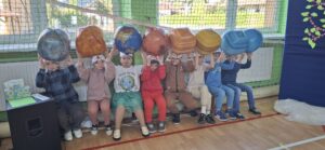 Grupa dzieci siedzi na ławce w kolorowych strojach nad głową trzymają modele planet