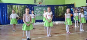„Grupa dzieci w kolorowych kostiumach tańczy na sali gimnastycznej podczas przedstawienia szkolnego zatytułowanego 'Moja Planeta'. Scenografia w tle przedstawia las, Ziemię oraz futurystyczną stację kosmiczną