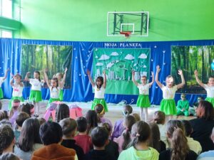 Grupa dzieci w kolorowych kostiumach tańczy na sali gimnastycznej podczas przedstawienia szkolnego zatytułowanego 'Moja Planeta'. Scenografia w tle przedstawia las, Ziemię oraz futurystyczną stację kosmiczną. Widzowie – inne dzieci – siedzą z przodu, obserwując występ