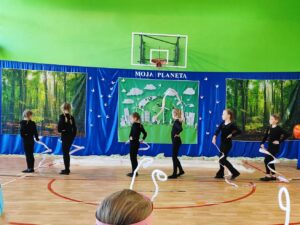 „Grupa dzieci w czarnych kostiumach tańczy na sali gimnastycznej podczas przedstawienia szkolnego zatytułowanego 'Moja Planeta'. Scenografia w tle przedstawia las, Ziemię oraz futurystyczną stację kosmiczną