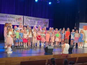 Grupa dzieci ubranych w kolorowe stroje stojących na scenie podczas konkursu wokalnego.