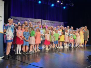 Grupa dzieci ubranych w kolorowe stroje stojących na scenie podczas konkursu wokalnego.