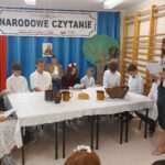 Baner: Narodowe Czytanie - Patronat Honorowy Pary Prezydenckiej. Po środku zdjęcie Jana Kochanowskiego. Pod nim siedzi uczniem ucharakteryzowany na Jana Kochanowskiego. Z lewej strony flaga biało-czerwona. Po prawej lipa z kartonu, pod nią dziewczynka na zielono i w wianku - przebrana za lipę. 10 uczennic i uczniów siedzi za stołem nakrytym białym obrusem. Na stole kosz z owocami, chleb i dzbanki. Obok stoi Pani Dyrektor, która czyta utwór Kochanowskiego.